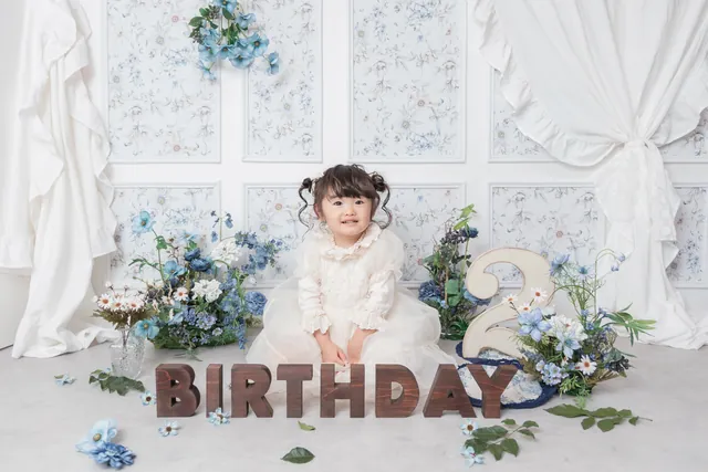 「BIRTHDAY」の文字と青い花のセットで、白いドレスの女の子が座っている写真