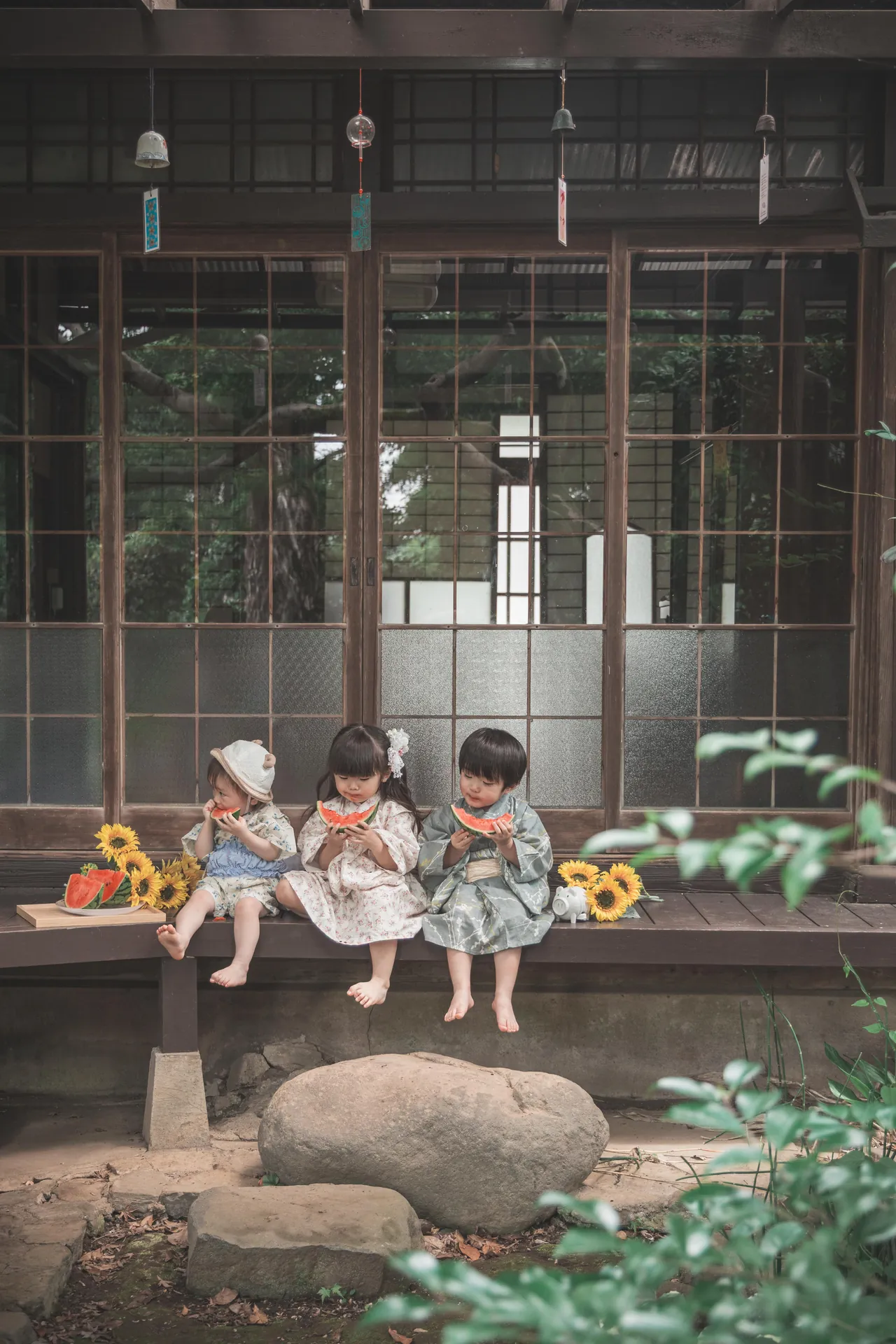 古い建物の縁側で、三人の子どもがすいかを食べている写真