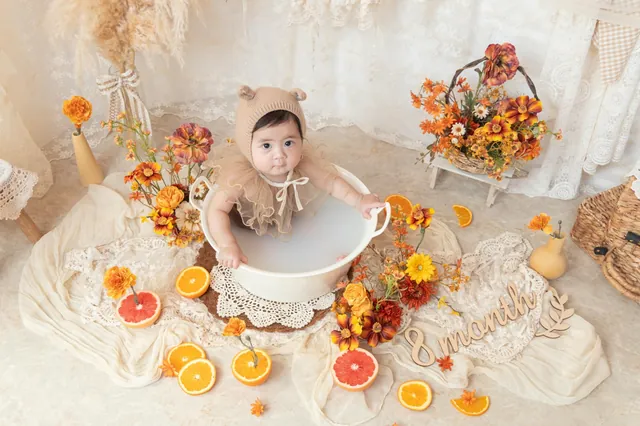 オレンジの花と果物に囲まれたミルクバスで、くまの耳の帽子をかぶった赤ちゃんがこちらを見上げている写真